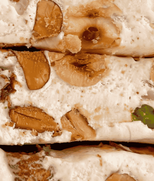 Design sans titre (17) Gros plan sur des tranches de nougat royal artisanal, richement garnies d'amandes, noisettes et pistaches, révélant une texture moelleuse.