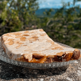 Tranche généreuse de nougat artisanal au caramel beurre salé et amandes entières, présentée en extérieur sur un plat en verre.
